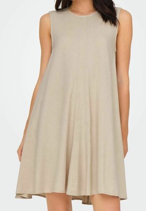 Femme portant une robe beige sans manches, coupe trapèze jusqu'aux genoux, avec un col rond, debout devant un fond blanc.