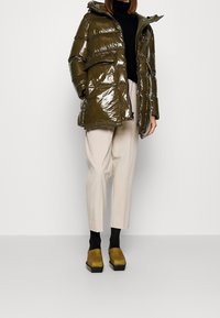 Chaqueta de plumas verde oliva brillante con capucha, cierre de cremallera y bolsillos frontales; combinada con pantalones crema y zapatos de plataforma amarillos.
