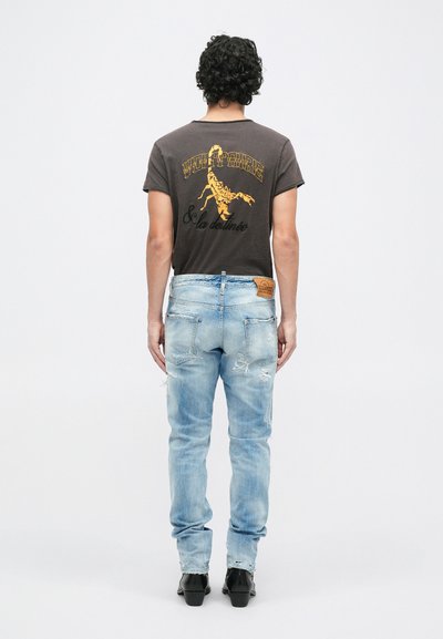 T-shirt gris avec un graphique de scorpion jaune, associé à un jean déchiré bleu clair. Modèle portant des chaussures noires, vu de dos.