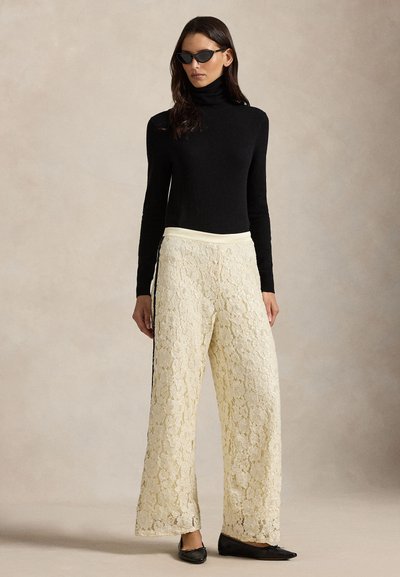 Svart polotröja, krämfärgade vida byxor i spets med texturerat mönster och svarta platta skor. Modellen bär kattögonformade solglasögon.