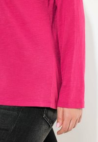 Langärmliges Shirt in leuchtendem Pink mit glatter Textur und abgerundetem Saum, kombiniert mit dunklen Denim-Jeans, bei denen eine Gesäßtasche sichtbar ist.
