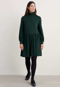 Zielona sukienka o długich rękawach z wysokim kołnierzem, marszczoną talią i rozkloszowanym dołem. Gładki materiał i jednolity kolor. Noszona z czarnymi rajstopami i butami.