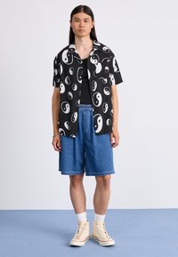 Chemise noire à manches courtes avec un motif yin-yang blanc, associée à un short en denim bleu et à des sneakers crème à tige haute avec des chaussettes blanches.