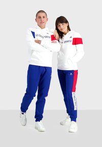 Des sweatshirts blancs avec des accents rouges et bleus, associés à des pantalons de survêtement bleus avec un imprimé logo blanc. Les deux portent des baskets blanches.