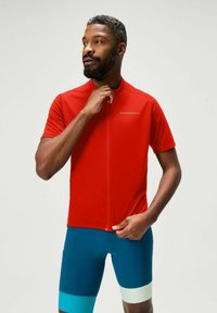 Rotes Fahrradtrikot mit Reißverschluss und kurzen Ärmeln, kombiniert mit blauen Fahrradhosen mit türkisfarbenen und weißen Akzenten. Glatter, sportlicher Schnitt.