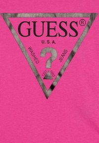 Roosa puuvillane T-särk, millel on must kolmnurga graafika koos tekstiga "GUESS U.S.A.", küsimärgi ja "WASHED JEANS" sildiga.
