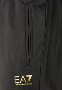 Pantaloni sportivi neri realizzati in tessuto testurizzato, con una tasca con zip verticale e il logo dorato EA7 Emporio Armani in basso a sinistra.