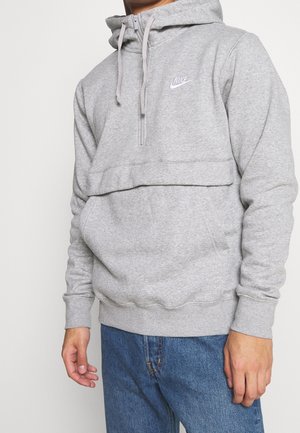 Mężczyzna w jasnoszarej bluzie Nike z kapturem, suwakiem z przodu i troczkami, w połączeniu z niebieskimi dżinsami, stoi na jednolitym tle.