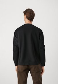 Felpa nera con scollo rotondo e maniche lunghe, caratterizzata da una superficie liscia e una sottile etichetta sul retro. Indossata con pantaloni marroni.