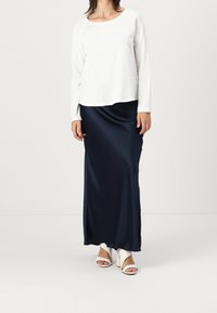 Top bianco a maniche lunghe e girocollo abbinato a una lunga gonna maxi in satin blu notte. Ai piedi, sono visibili dei sandali bianchi con cinturini.