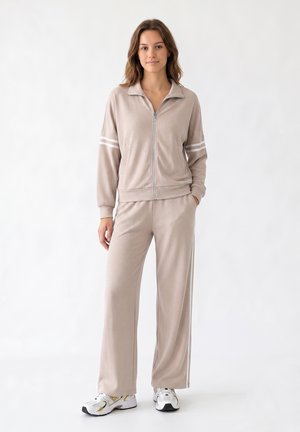 Jeune femme debout portant un survêtement beige avec des bandes blanches sur les manches et le pantalon, associé à des chaussures de course blanches et dorées.