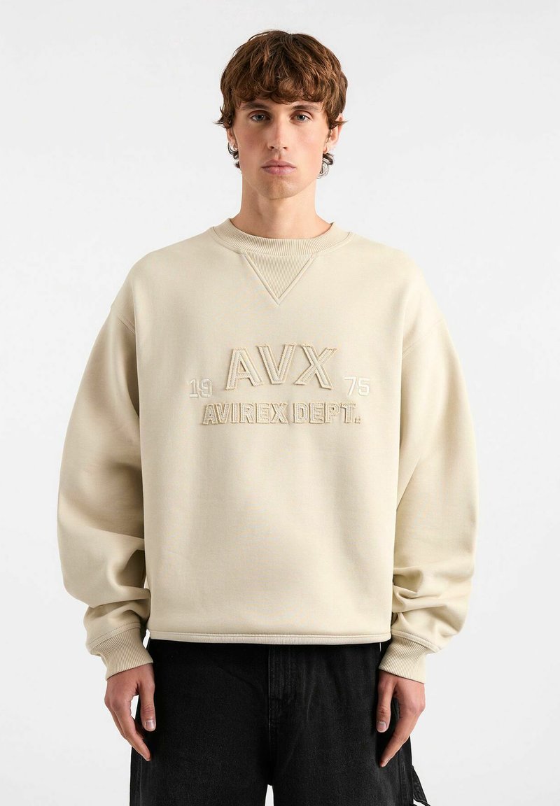 Felpa beige oversize con scollo a girocollo a coste, presenta il testo in rilievo "AVX 1975 AVIREX DEPT." nella parte anteriore e maniche a vestibilità ampia.