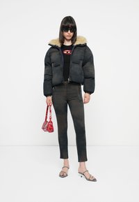 Black puffer jacket with fur collar, fitted black top, dark jeans, silver sandals, and a red patterned handbag. Model poses against a plain background.