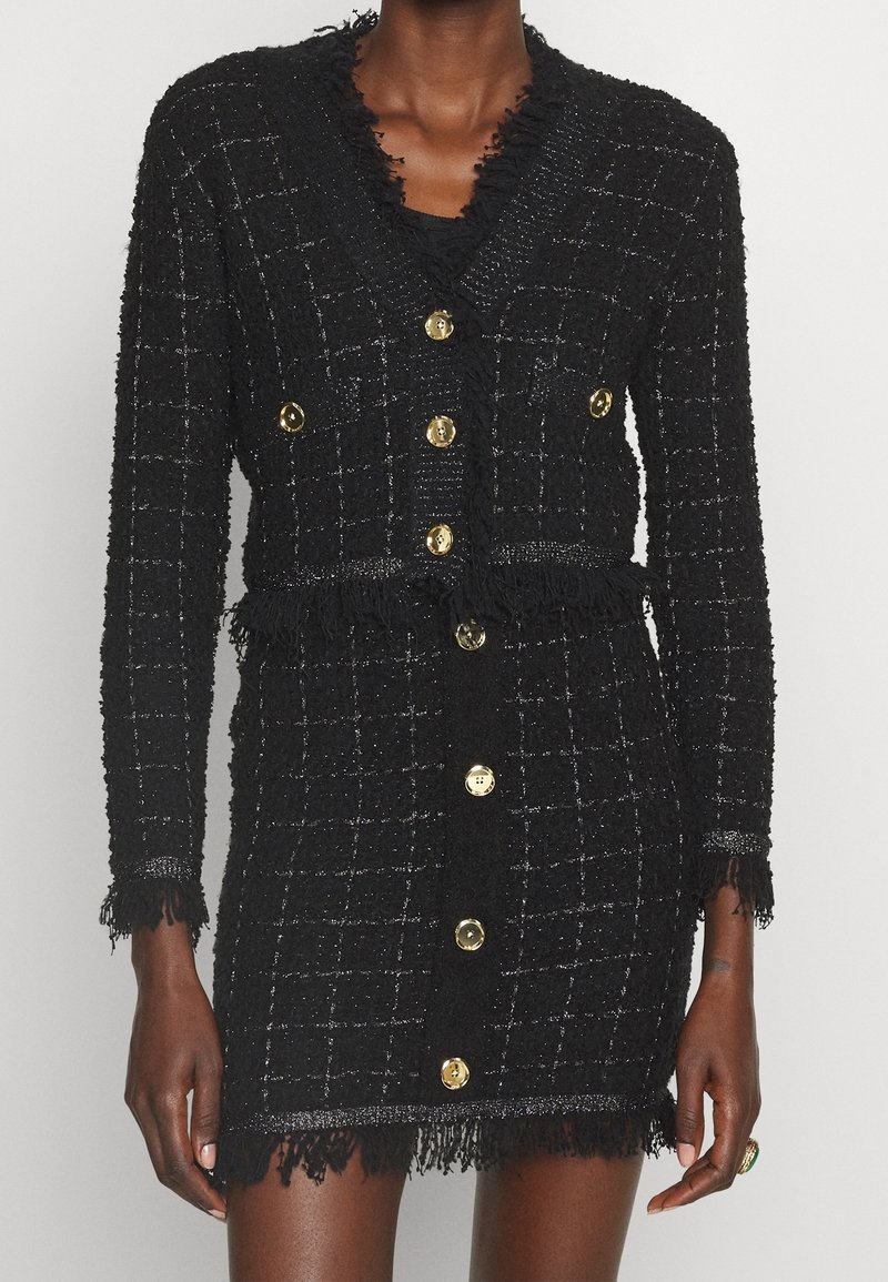 Robe en maille texturée noire avec des boutons dorés, bordure à franges sur le col, les poignets, la taille et l'ourlet, portée par une personne debout à la peau foncée.