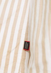 Pruhovaná tkanina v béžové a bílé barvě s texturovaným povrchem. Obsahuje štítek Scotch & Soda s černými a červenými akcenty.