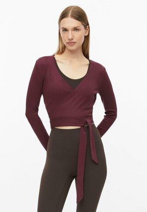 Femme portant un haut à manches longues style cache-cœur bordeaux par-dessus un soutien-gorge de sport noir et des leggings sombres taille haute, debout sur fond blanc.