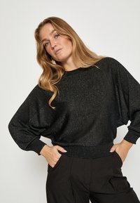 Black, sparkly knit sweater with a loose fit, wide sleeves, and ribbed cuffs. Paired with black pants, displaying an elegant, relaxed silhouette.