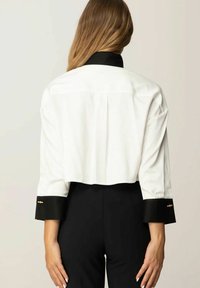Blusa bianca corta con colletto e polsini neri, caratterizzata da una texture liscia e dettagli hardware dorati sul polsino. Vista posteriore.