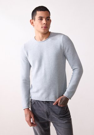 Ljusblå stickad tröja med rund halsringning och långa ärmar, tillsammans med grå jeans. Texturen verkar mjuk med synliga ribbadeta detaljer.