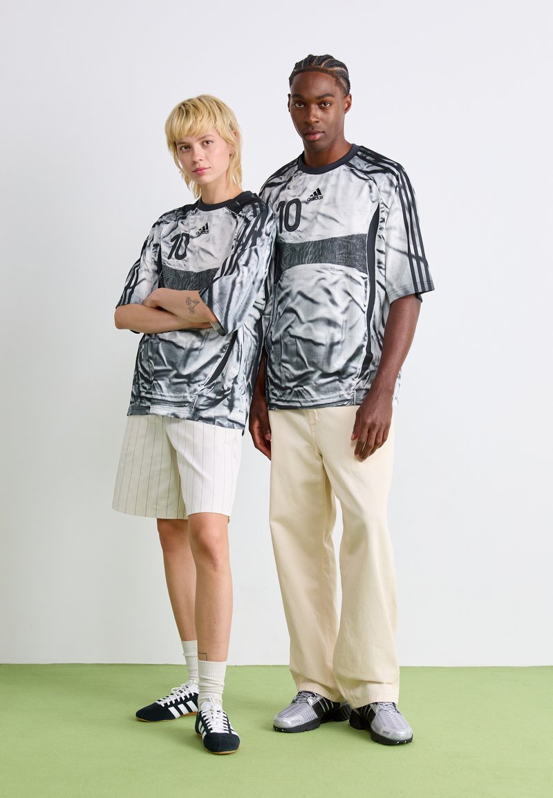 Dos modelos de pie sobre un suelo verde contra una pared blanca, vistiendo camisetas gris Adidas a juego con el número 10, uno con pantalones cortos a rayas y el otro con pantalones crema.