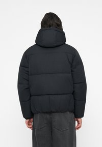 Jachetă puf din material negru, cu glugă, având un design matlasat, o siluetă pufoasă și un tiv elastic. Vederea din spate arată textură netedă a materialului.