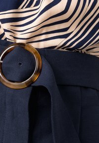 Vêtement ceinturé en tissu bleu marine, taille cintrée, et une grande boucle en écaille de tortue. Un haut crème à rayures bleu marine est partiellement visible.