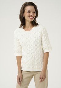 Pull en tricot blanc avec un motif torsadé texturé agrémenté de perles et des manches trois-quarts, associé à un pantalon beige clair.