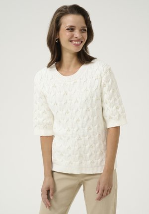 Pull en tricot blanc avec un motif torsadé texturé agrémenté de perles et des manches trois-quarts, associé à un pantalon beige clair.