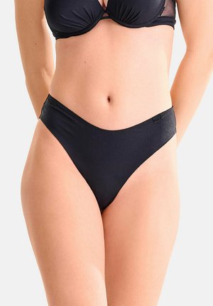 Femme portant un haut de bikini noir et un bas de bikini taille haute, debout les mains derrière le dos sur un fond uni.