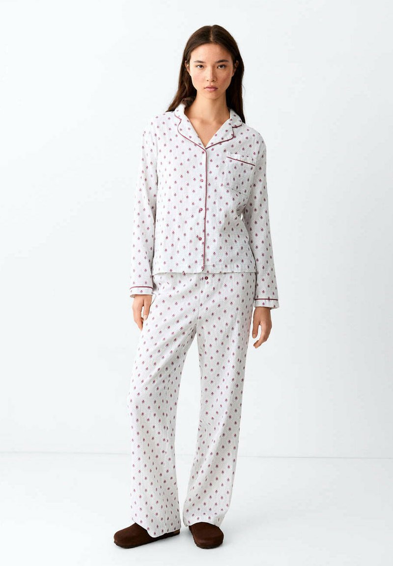 Femme portant un pyjama blanc à motifs à manches longues avec des détails rouges et des chaussons marron, debout devant un fond blanc uni.