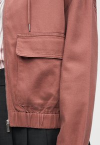 Κοντινό πλάνο σε ροζ μπουφάν με τσέπη με καπάκι, φερμουάρ και ελαστική μέση, φορεμένο πάνω από μαύρο παντελόνι και ριγέ πουκάμισο.