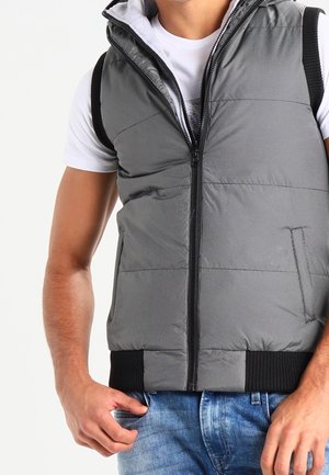 Man draagt een grijs gewatteerd mouwloos vest met zwarte bies over een wit T-shirt en blauwe spijkerbroek, hand rustend bij de zak.