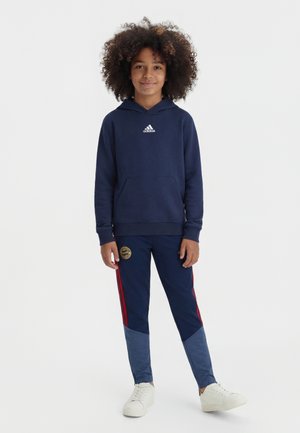 Barn med krøllet hår har en mørkeblå Adidas hættetrøje og matchende træningsbukser med røde striber på, stående med hænderne afslappet ved siden.
