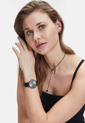 Schwarze Uhr mit einem runden, minimalistischen Zifferblatt und strukturiertem Lederarmband. Die Trägerin trägt eine geschichtete schwarze Kettenhalskette und hat lange, fließende Haare.