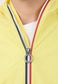 Geel lichtgewicht jack met een opvallende ritssluiting met een zilveren trekker, geflankeerd door rode en donkerblauwe accenten. Soepele textuur.