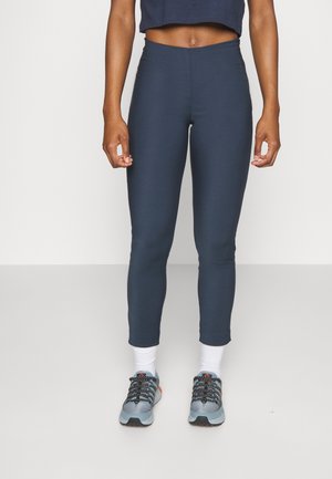 Osoba nosząca granatowe skrócone legginsy, białe skarpetki i szare buty sportowe, stojąca na jednolitym białym tle.