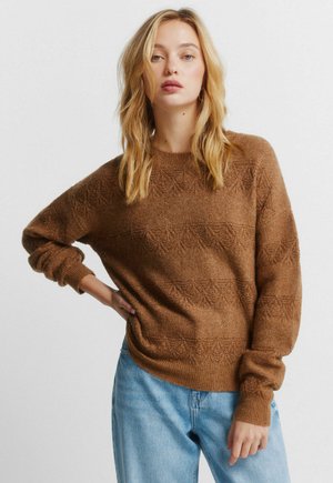 Brauner Strickpullover mit texturierten Blattmustern, rundem Ausschnitt und gerippten Bündchen. Kombiniert mit hellblauen Denim-Jeans.