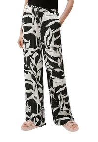 Pantalon large noir et blanc avec un motif de feuilles, tissu léger, taille élastique et poches latérales. Porté avec des sandales rose clair.