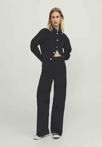 Casaco de ganga preto com um design cropped e grandes botões brancos, combinado com calças largas de ganga preta. Tecido texturizado. Ajuste casual.