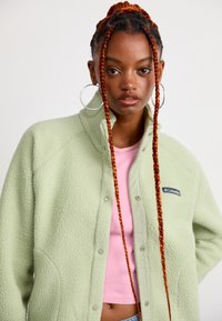 Jeune femme aux longues tresses rouges portant de grandes boucles d'oreilles en anneau, une veste en polaire Columbia vert clair et un crop top rose.