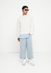 Pulover alb tricotat cu mâneci lungi, suprapus peste o cămașă albastră cu dungi. Pantaloni din denim bleu, evazați, și adidași albi cu accente gri.