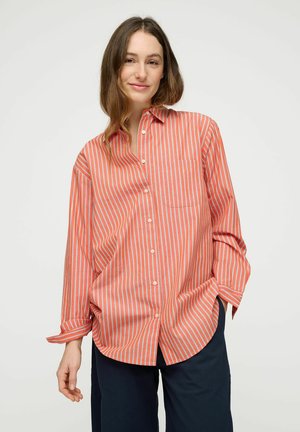 Kobieta o długich brązowych włosach, ubrana w oversize'ową pomarańczowo-białą koszulę w paski na guziki oraz ciemne spodnie, stojąca na jednolitym tle.