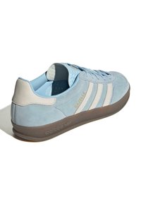 Gaiši zili zamšveida sporta apavi ar baltiem ādas svītrām, zelta logotipu, mīksto apkakli un gumijas zoli. Iespējas ietver noapaļotu purngalu un mīkstinātu iekšpusi.