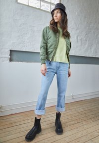 Zelena bomber jakna, svetlo zelena majica, svetlo plave zarolane farmerke i crne čizme do članaka. Pod od drveta i pozadina od teksturisanog belog zida.