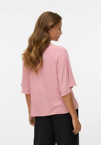 Donna con lunghi capelli ondulati che indossa una maglietta corta rosa morbida e pantaloni neri, rivolta di lato su uno sfondo bianco semplice.