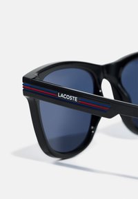 Mustat aurinkolasit, joissa on sinisiä ja punaisia raidallisia yksityiskohtia varsissa. Ne sisältävät tummat sävytetyt linssit ja geometrisen muotoilun. "LACOSTE"-logo esillä.