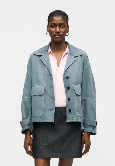 Femme portant une veste bleue ample sur une chemise boutonnée rose clair et une jupe gris foncé, debout devant un fond uni.