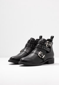 Botas de cuero negras hasta el tobillo con hebillas plateadas y correas con remaches, tacones bajos y tiradores sobre una superficie blanca.
