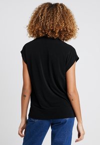 Person med lockigt hår står vänd bort, iklädd en svart ärmlös topp och blå jeans, med små tatueringar på båda armarna.