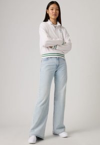 White zip-up jacket with green stripes, high collar, paired with light blue wide-leg jeans and white sneakers, emphasizing casual style and comfort.